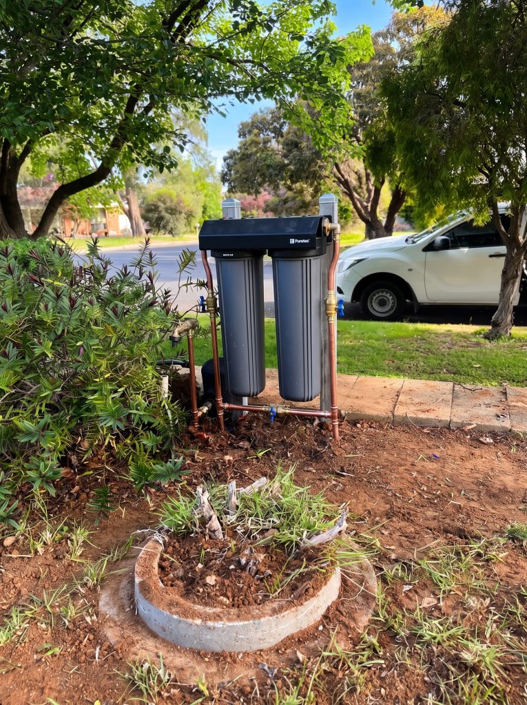Twin-cartridge water filtration system installed beside a garden bed outside a home