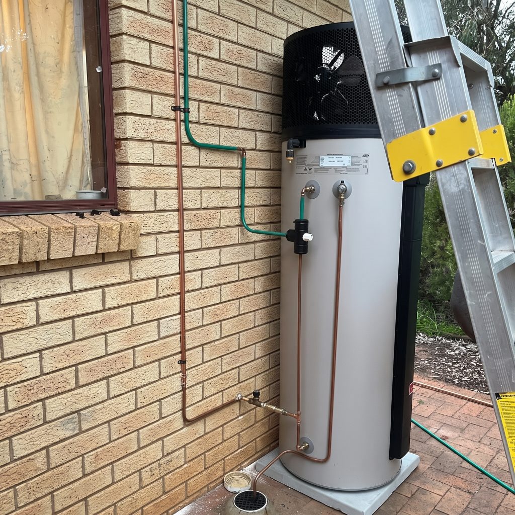 Heat pump hot water unit installed against a brick wall with a ladder alongside